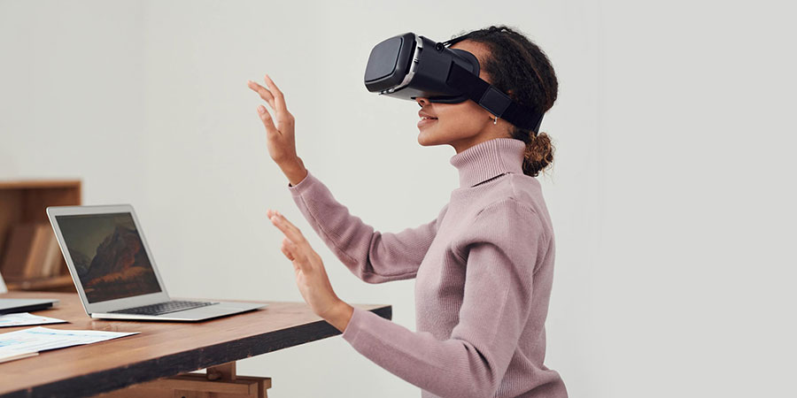 a woman in light purple long sleeve turtle neck sweater sitting in front of a laptop on top of a table while using a virtual reality goggles