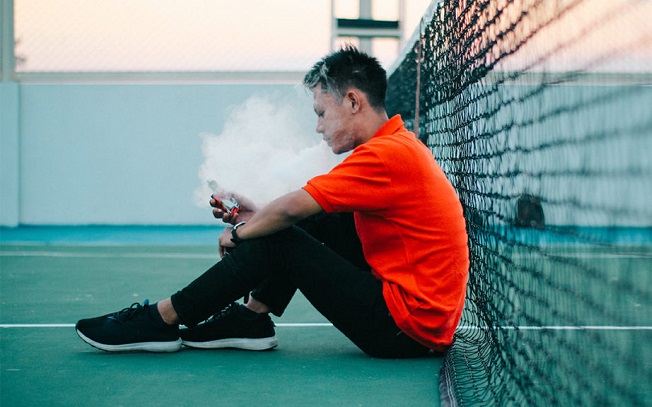 Side View Of Young Man Smoking Electronic Cigarette While Sitting On Tennis Court